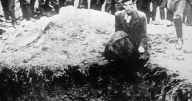 The heartbreaking photograph shows a German soldier aiming a gun at a man kneeling beside a mass grave while he looks into the distance