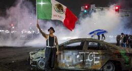 A protester waves the Mexican flag beside the wreckage of a burnt car sprayed with graffiti against US Immigration and Customs Enforcement (ICE), as law enforcement clashes with demonstrators during a protest following federal immigration operations, in the Compton neighborhood of Los Angeles, California on June 7, 2025