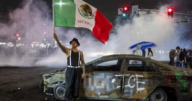 A protester waves the Mexican flag beside the wreckage of a burnt car sprayed with graffiti against US Immigration and Customs Enforcement (ICE), as law enforcement clashes with demonstrators during a protest following federal immigration operations, in the Compton neighborhood of Los Angeles, California on June 7, 2025