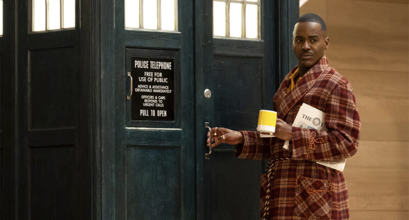 Ncuti Gatwa wearing a tartan dressing gown and holding a cup of tea standing by the TARDIS